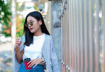 A beautiful teenage girl wearing a white t-shirt, jeans and dark glasses holds a red teapot glass against the wall, looking cool and charming.