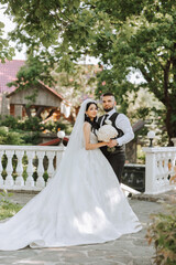 Fototapeta premium A handsome groom embraces his bride in a lush white dress and smiles in a beautiful outdoor setting. Under the open sky. High quality photo. A newlywed couple poses together on a sunny summer day.
