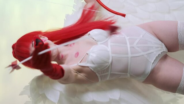 Beautiful girl with red hair, in a white bodysuit, red heart-shaped glasses, red gloves. White wings on the back, traces of kisses on the body. Bow and arrows in hand. Sexy cupid. Vertical video
