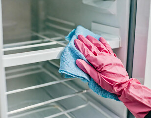 Young adult woman hand in pink rubber protective glove wiping inside plastic box of white freezer with dry blue rag. Closeup. Front view.