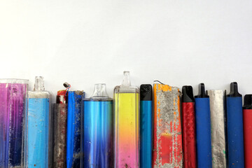 Multi-coloured discarded electronic cigarette vapes lined up on a white paper background. These are...