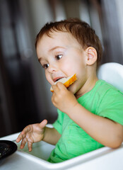 Little boy sits in high chair and eats piece of orange