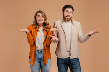 Clueless and puzzled stylish male and female standing at beige wall, having confused looks, shrugging shoulders isolated on background