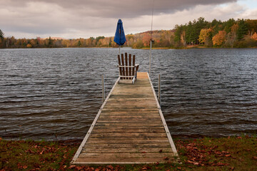 Wooden dock over the lake and an empty Adirondack chair near a shadow and a fishing rod in an...