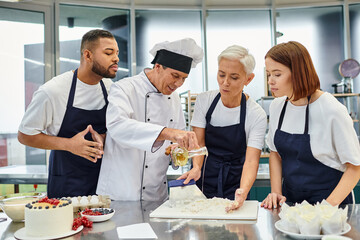 joyful multiracial chefs in blue aprons watching chief cook pouring oil on dough, confectionery