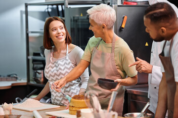 jolly female students in aprons smiling at each other next to their african american friend and chef