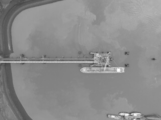 top down view on a inland shipping bulk carrier docked moored at a loading and unloading terminal pumping liquid of gas products into containers on land.