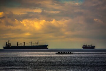 Silhouettes of two container ships contrasted with a silhouette of Indians paddling a long canoe, Vancouver harbor, Canada