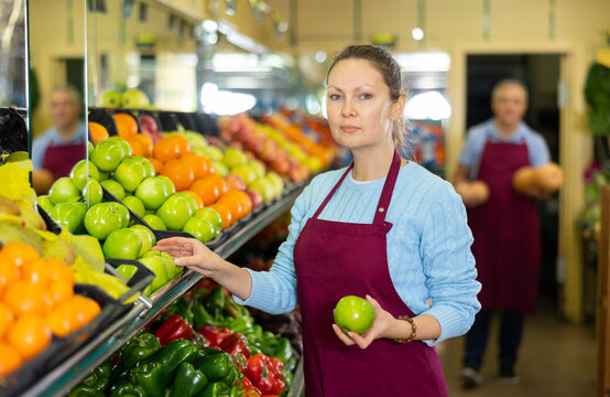 Skillful female merchandiser in uniform checking green apples before selling in grocery supermarket