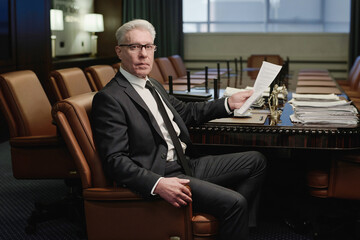 Mature experienced lawyer posing in armchair at table in conference room looking through papers