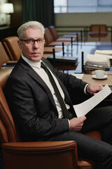 Senior businessman sitting alone in office boardroom holding documents and looking at camera