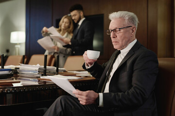 Mature lawyer sitting and reading document while drinking coffee, his colleagues working with papers