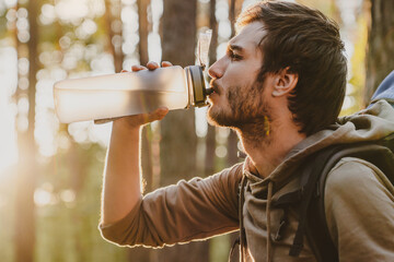 Handsome young guy drinking water while walking and traveling by pine forest with copy space. Rehydration with cold beverage during hiking backpacking alone in woods