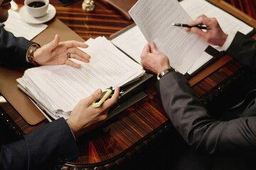 Two unrecognizable business partners discussing documents while sitting at wooden desk in boardroom
