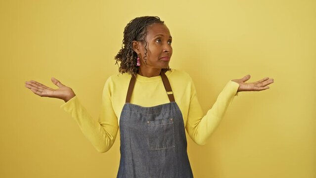 Clueless and confused, a beautiful middle-aged african american woman, apron-clad, stands with raised hands against an isolated yellow background, capturing a perfect picture of doubt.