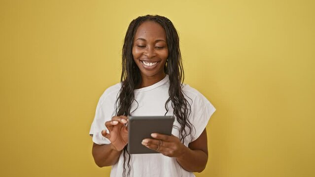 A smiling woman with curly hair holds a tablet in front of a plain yellow wall, depicting casual technology use.