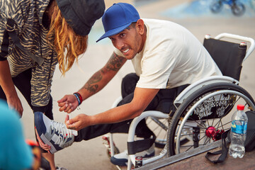 athlete man with disability in wheelchair smiling while fasten sneakers with help at skatepark	