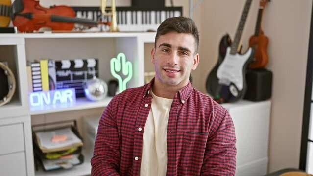 Upbeat portrait, confident, smiling young hispanic male musician sitting indoors in music studio, ready for melodic performance