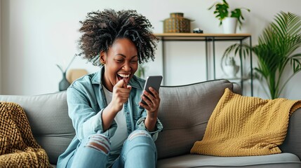 Happy young black woman sitting on sofa at home with phone in her hand. - obrazy, fototapety, plakaty