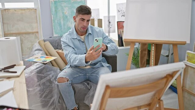 Young, confident hispanic man artist happily making photo via smartphone to paint on canvas in art studio