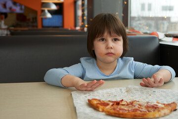A child with his cheeks smeared with sauce pushes half a pizza away from him. The interior of a...