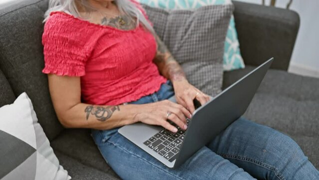 Focused grey-haired middle-age woman using laptop on the living room sofa at home