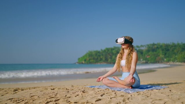 Woman in yoga pose meditates with VR glasses on sandy beach. Virtual retreat experience, practices mindfulness beside sea. Tech meets zen, wellness in nature. Slow motion.