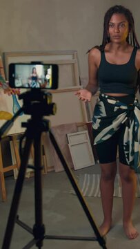 Vertical full length shot of backstage of young Black woman filming herself on phone using tripod and introducing on camera then showing dance moves at home
