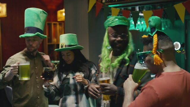 Medium footage of diverse adult friends wearing green hats dancing and clinking glasses of beer