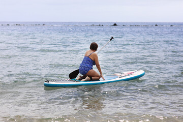 Middle aged woman in swimsuit sits on inflatable SUP board and paddling at calm ocean water. Active female enjoys does yoga, stretching or trains on paddle board at summer vacation. Healthy lifestyle