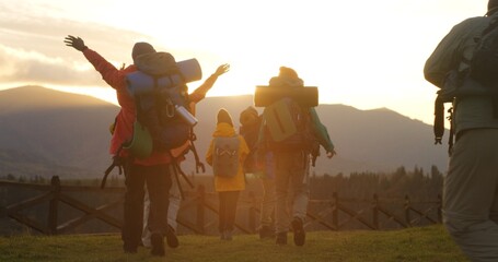 Diverse hikers with backpacks and trekking poles go hiking in mountains. Happy tourist family or hiking buddies during vacation road trip. Sunset and mountain landscapes in background. Slow motion.