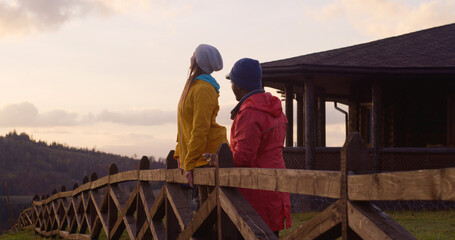 Multiethnic couple stands near wooden fence, looks at beautiful sunset and mountain landscapes enjoying nature during trip. Tourist family rest after hike or expedition in the mountains. Slow motion.