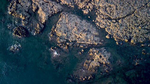 Top-down aerial view over flat rocks in blue pacific ocean during sunset