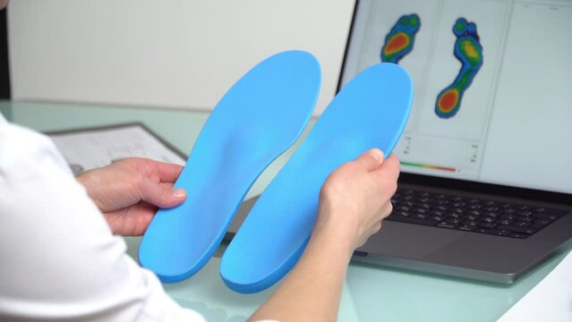Female doctor orthopedist  holding custom made insoles in a clinic in front of the laptop with a test feet picture.