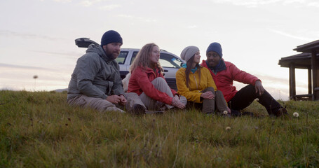 Group of multiethnic tourists sit on the grass at the hilltop and talk. Travelers or hiking friends enjoy nature during trip to the mountains. Stylish wooden cottage, car and dog in the background.