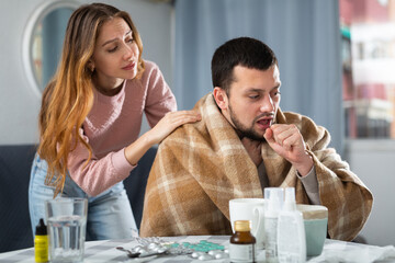 Young loving woman taking care of sick husband suffering from flu sitting wrapped in blanket in living room..