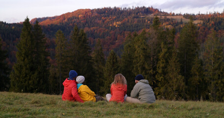Multiethnic couples sit on the grass on the top of the hill, look at sunset and amazing mountains scenery. Group of tourists or hiking friends enjoy beautiful nature during trip. Outdoor enthusiasts.
