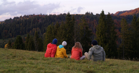 Multiethnic couples sit on the grass on the top of the hill, look at sunset and amazing mountains scenery. Group of tourists or hiking friends enjoy beautiful nature during trip. Outdoor enthusiasts.