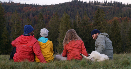 Diverse tourists or hiking buddies sit on the grass at the hilltop and pet dog. Group of travelers talk and enjoy nature during trip. Sunset and amazing mountain scenery. Outdoor exploration concept.