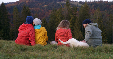 Diverse tourists or hiking buddies sit on the grass at the hilltop and pet dog. Group of travelers talk and enjoy nature during trip. Sunset and amazing mountain scenery. Outdoor exploration concept.