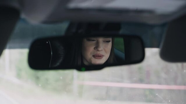 The girl is riding in the passenger seat of the car and talking to the driver. Her face is reflected in the rearview mirror.