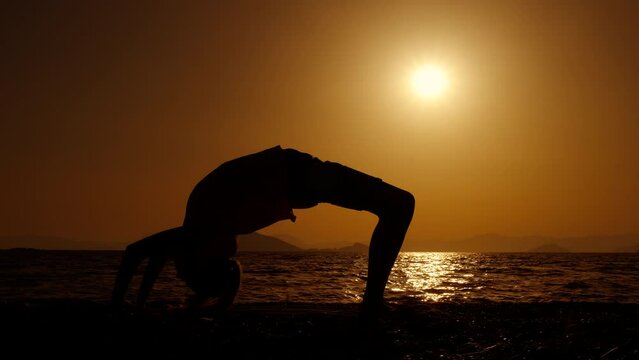 Teen silhouette doing yoga gymnastics. A sporty teen silhouette doing stretching gymnastic pose on the nightfall beach in summer.