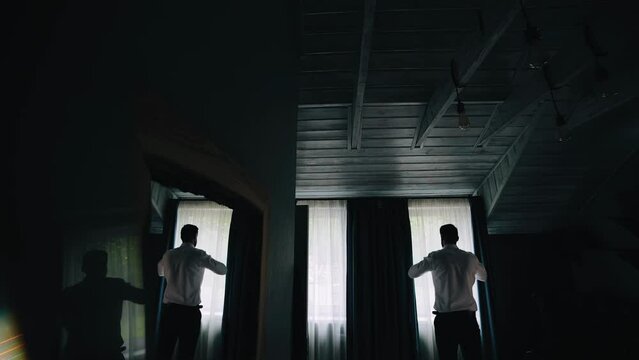 Shooting from the back of a man who stands in front of the window and gets dressed. His silhouette is visible in a dark room against a light window