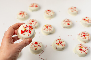 Hand Holding Decorated Valentine Cookie Dipped in White Chocolate and Decorated with Red, White, and Gold Sprinkles; More Cookies Out Of Focus in Background on White Parchment Paper