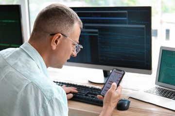 Fototapeta na wymiar Mature male programmer with mobile phone working in light office