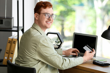 Handsome young man with credit card and modern laptop sitting at table while shopping online