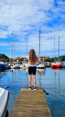 a young beautiful girl with long hair in a white shirt stands with her back to the viewer on a pier with yachts against the backdrop of the sea with her arms open, embracing the whole world and this w