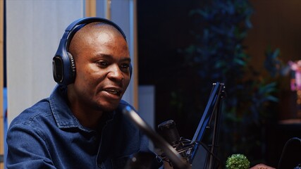 African american man doing podcast in studio, wearing headphones and talking on professional...