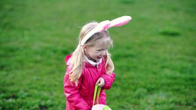 portrait of a small cute blond girl wearing Easter bunny ears on Easter egg hunt game in park on green grass. Easter egg hunt outdoors, copy space 