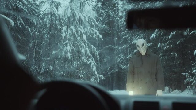 Curious alien in trench coat standing on the road in snowy forest, tilting head and staring at female driver sitting in the car. View through windshield